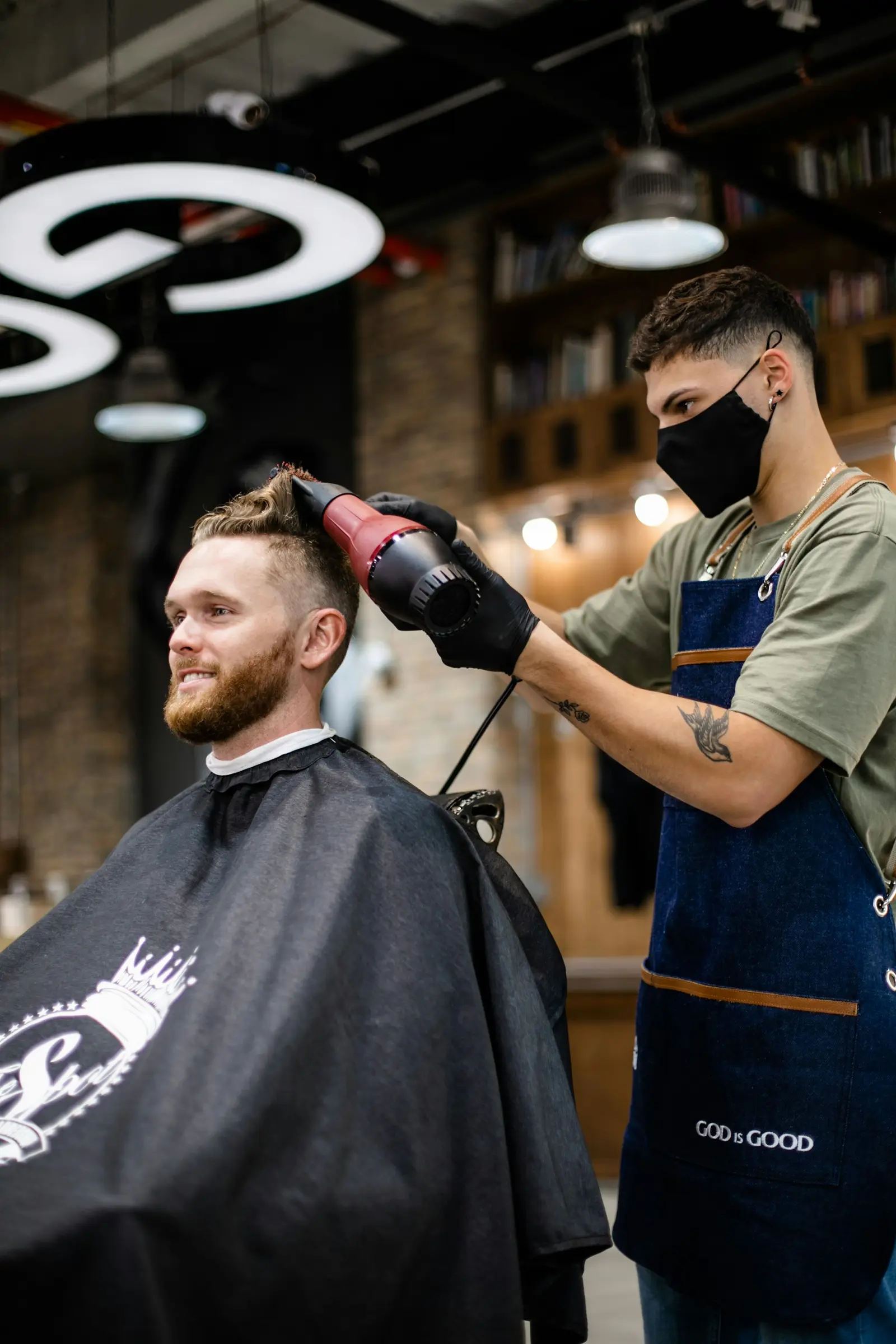 Barber in a face mask and gloves blow-drying a client's hair
