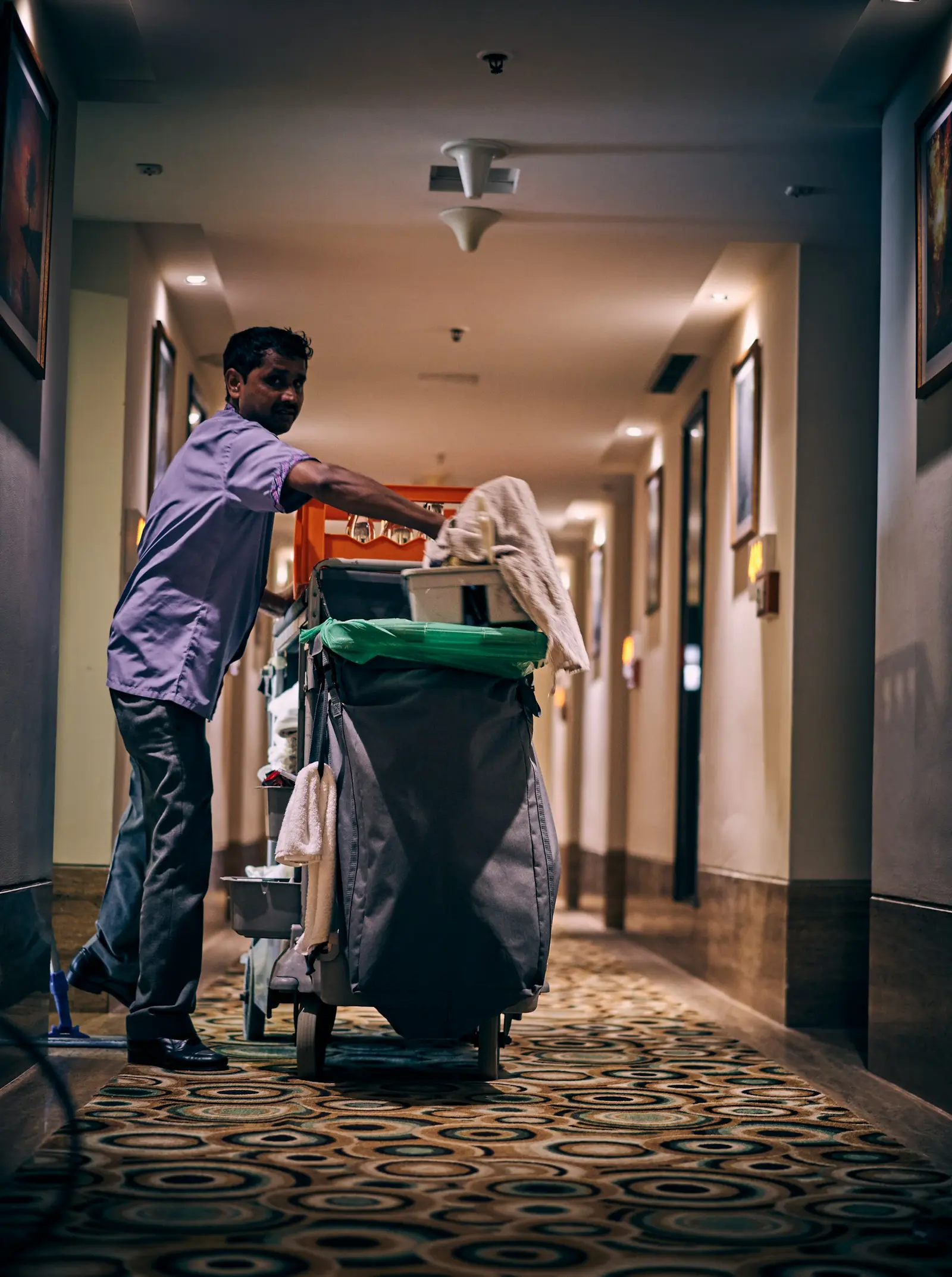 Housekeeper in a face mask pushing a cleaning cart along a hotel corridor